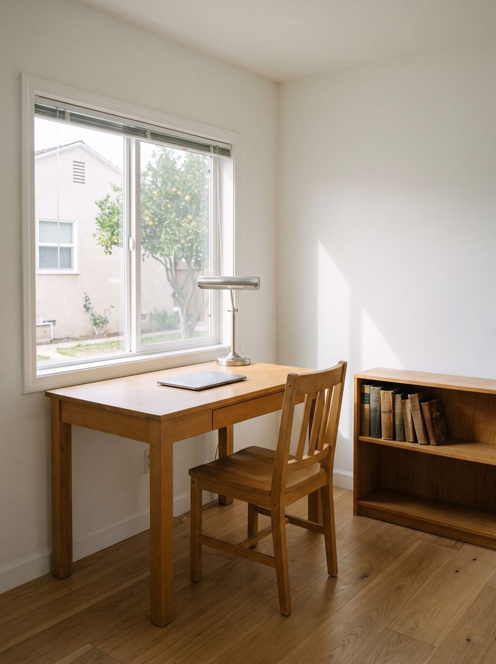 A desk under a window looking across a yard to the back of a stucco house